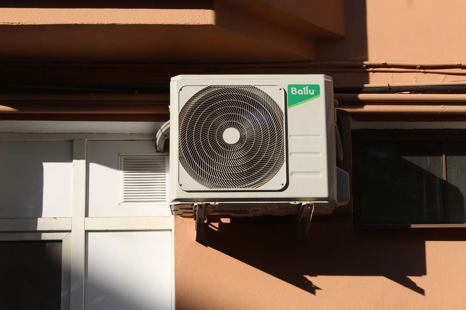 White outdoor air-conditioning condenser unit mounted on a house exterior wall in sunlight